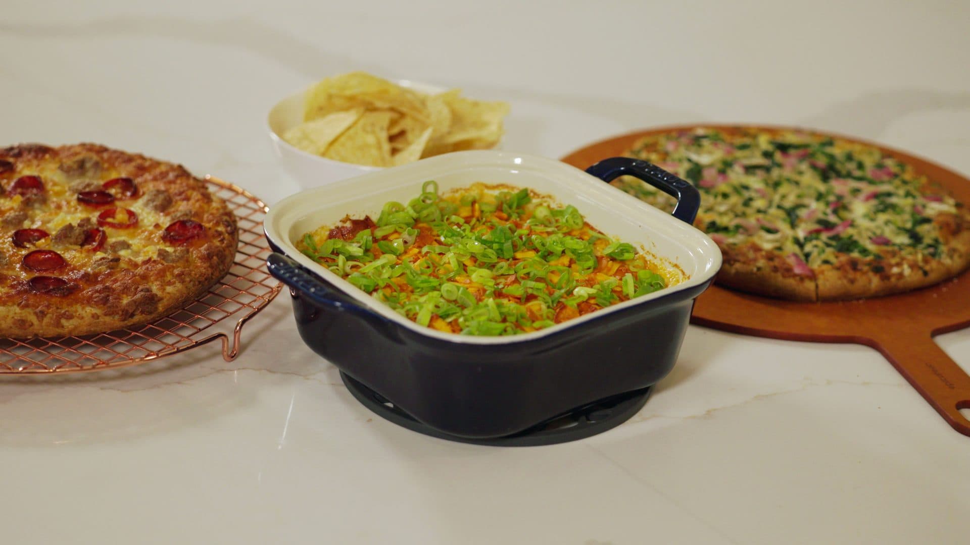 Buffalo chicken dip topped with sliced scallions in a navy baking dish, between two Real Dough pizzas and a bowl of tortilla chips
