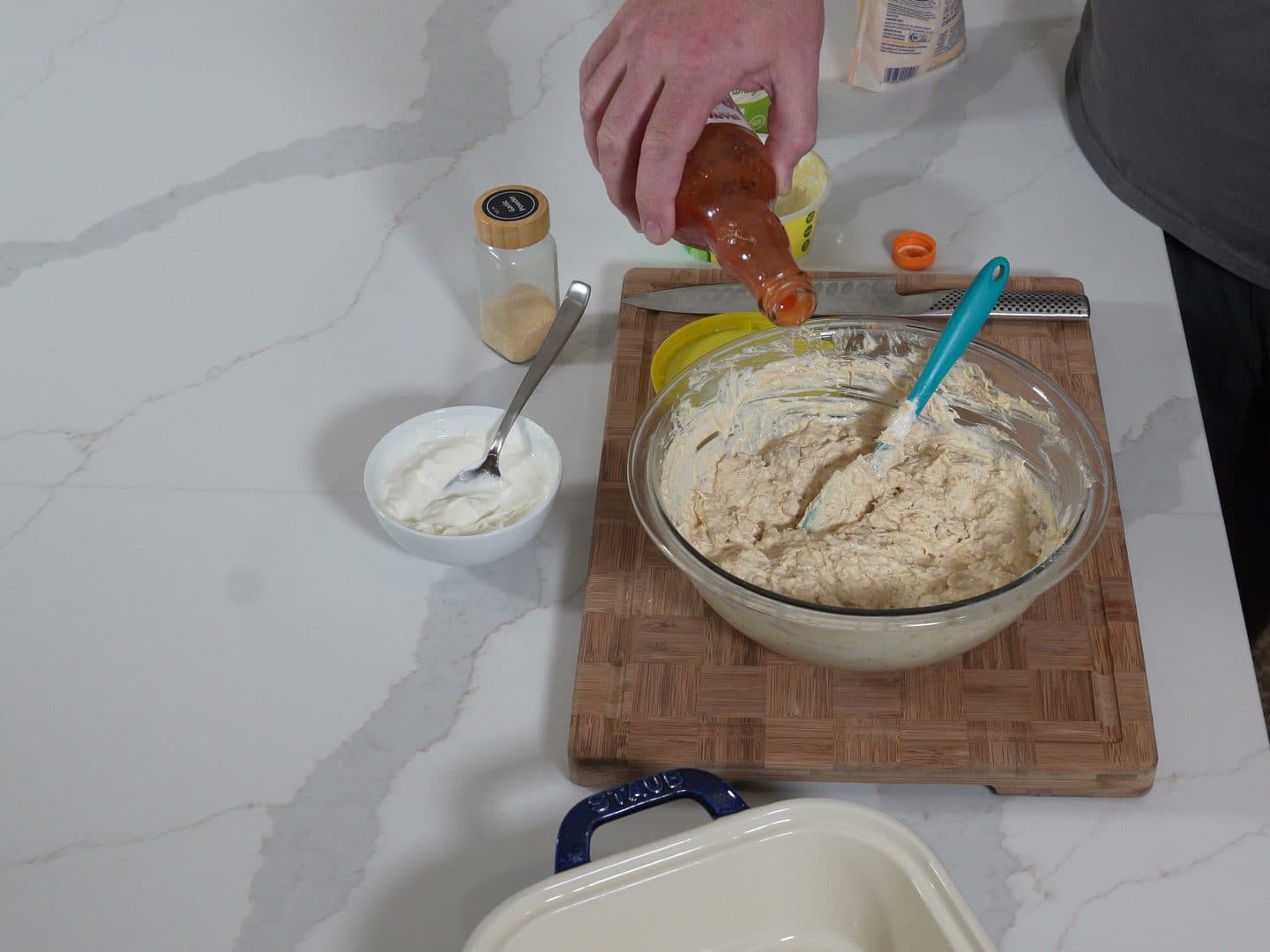 Overhead view of Frank's RedHot being poured into a glass bowl of creamy shredded chicken dip base on a wooden cutting board
