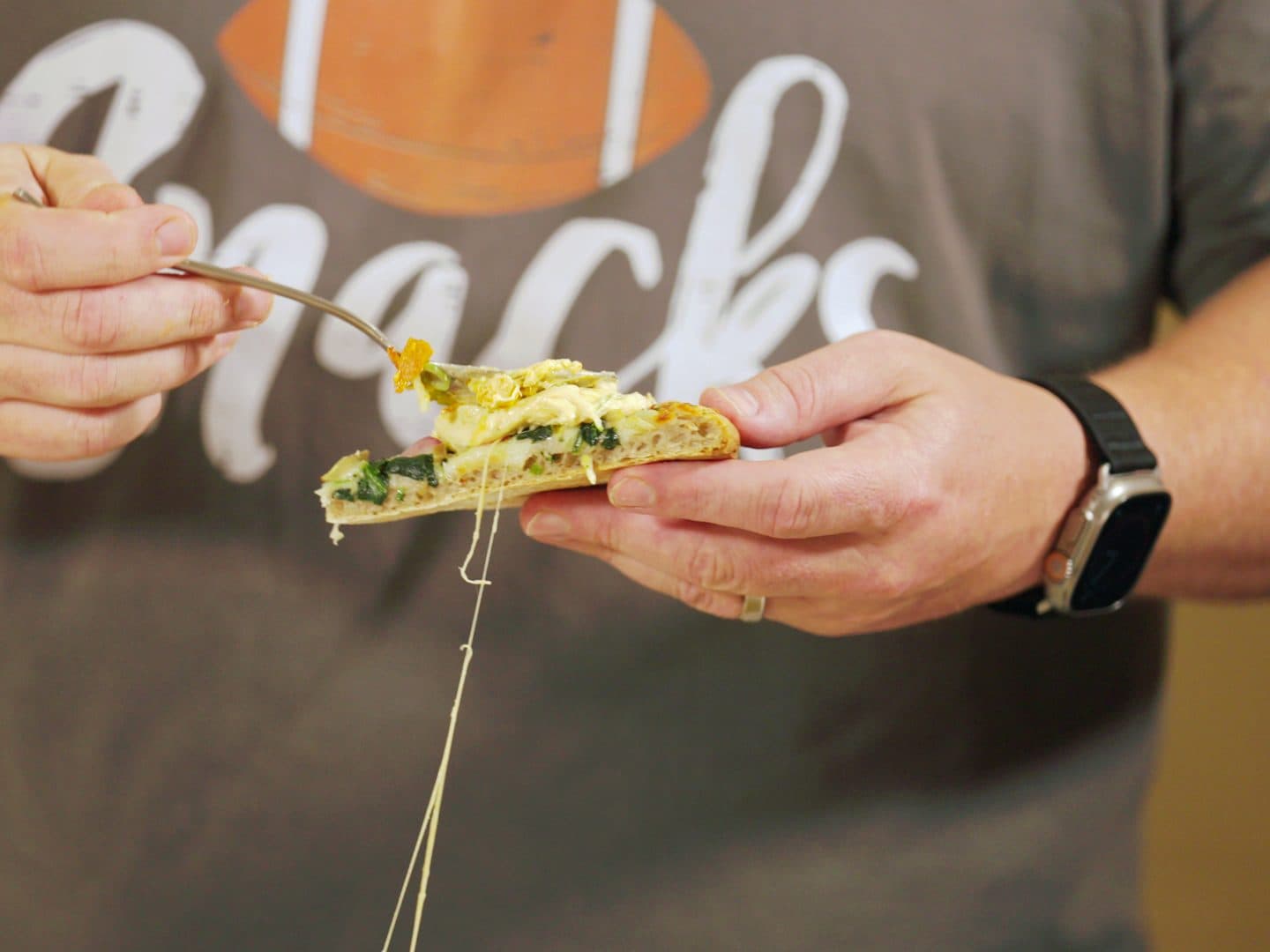 A spoonful of buffalo chicken dip going onto a slice of Okie Dokie Artichokie, with a long cheese pull hanging from the sourdough crust