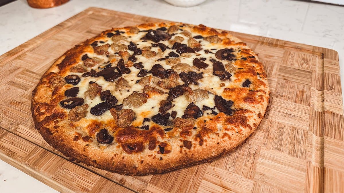 A baked Lost in the Sausage pizza on a wooden cutting board, sausage and mushrooms over bubbling white cheese and a deep golden sourdough crust
