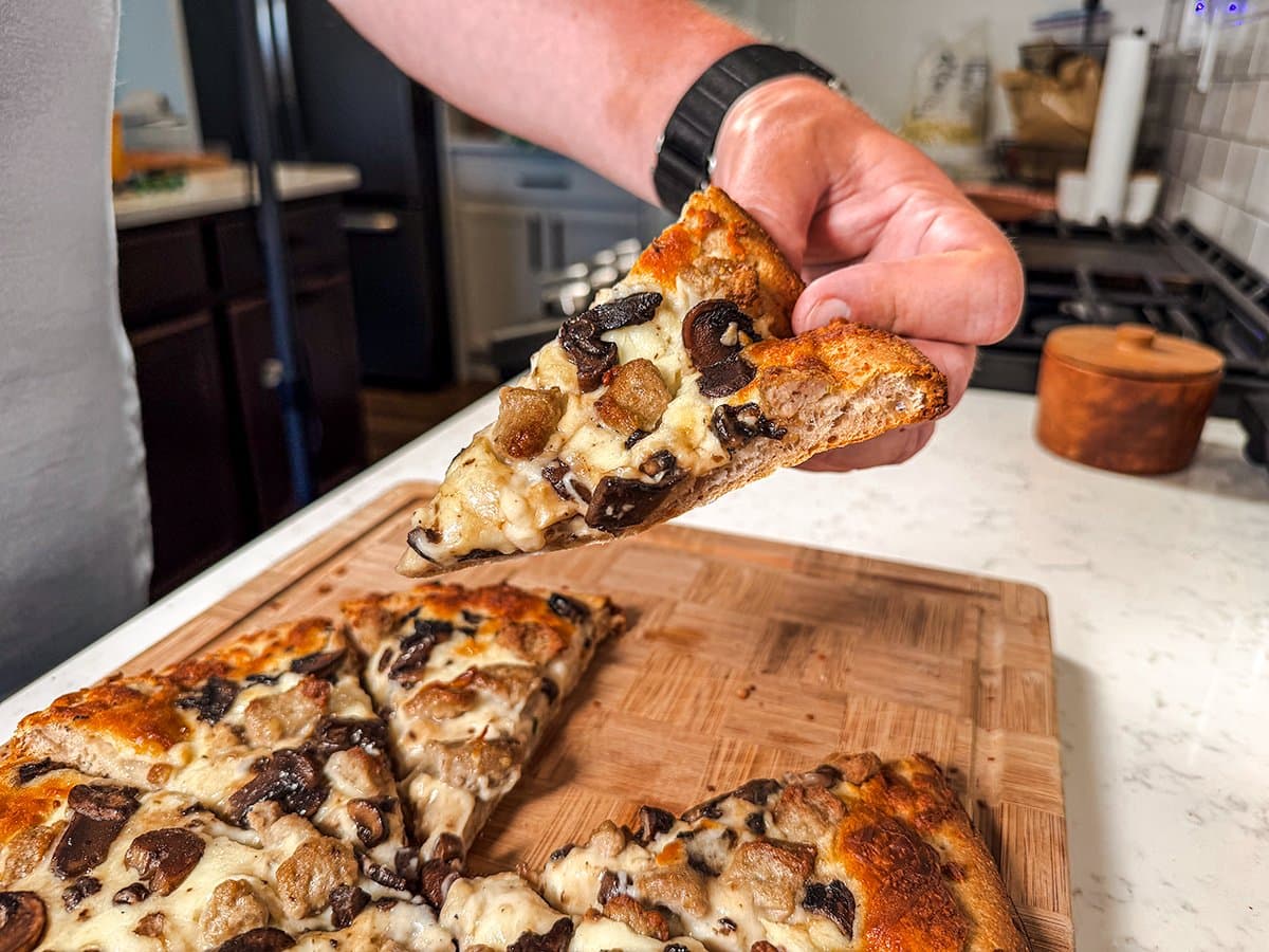 A hand holding up a slice of Lost in the Sausage over a cutting board, showing browned cheese and a crisp, golden sourdough edge