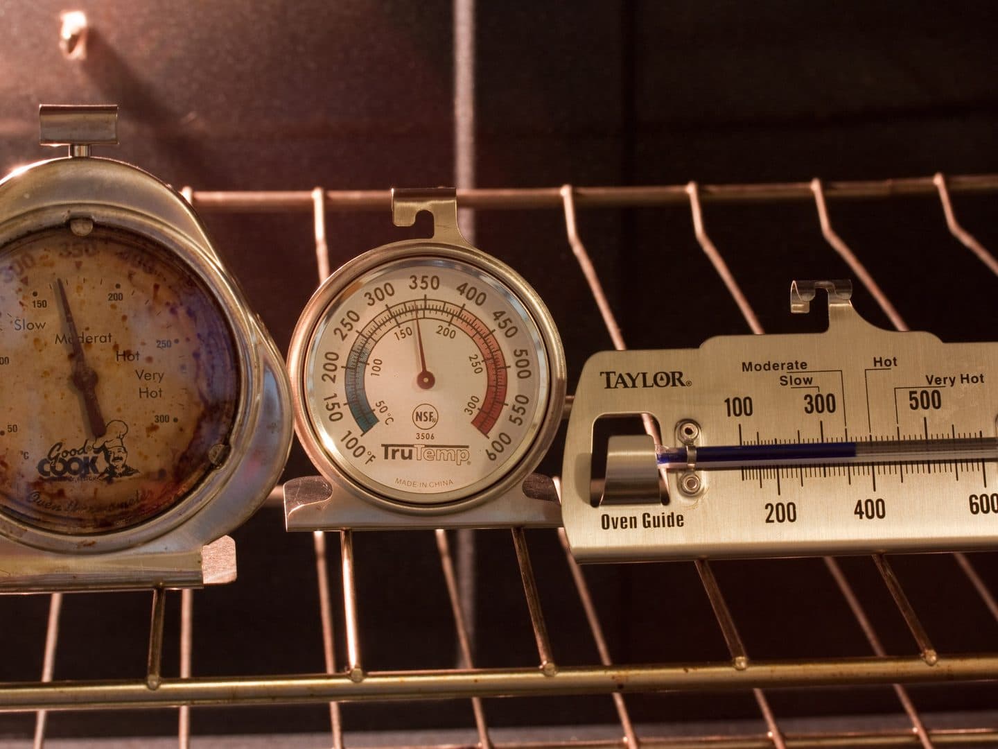 Three dial oven thermometers hanging side by side on an oven rack, each pointing at a different temperature