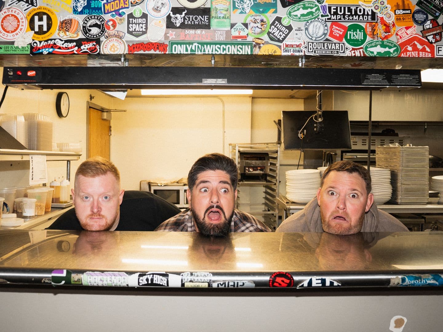 Real Dough co-founders Andrew Miller, Sam Emery and Cameron Whyte peeking over a restaurant kitchen pass, Sam mid-gasp